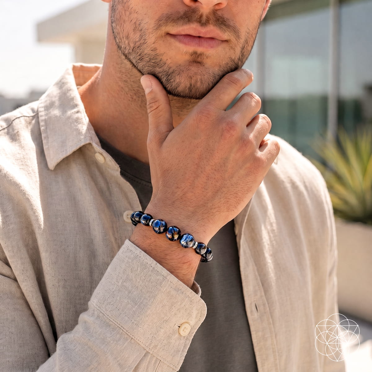Man wearing a blue pietersite and dark obsidian bead bracelet with a beige shirt, his hand resting beneath his chin.
