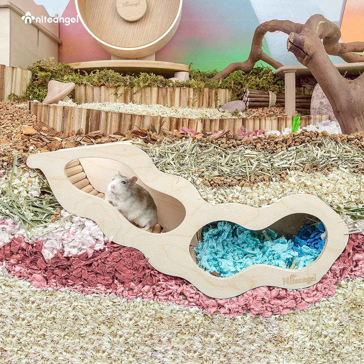A small gray hamster sits inside a wavy, light natural wood hide house with two openings, one containing blue paper bedding. The scene is a fully decorated hamster enclosure filled with wood shavings, hay, branches, and enrichment toys.