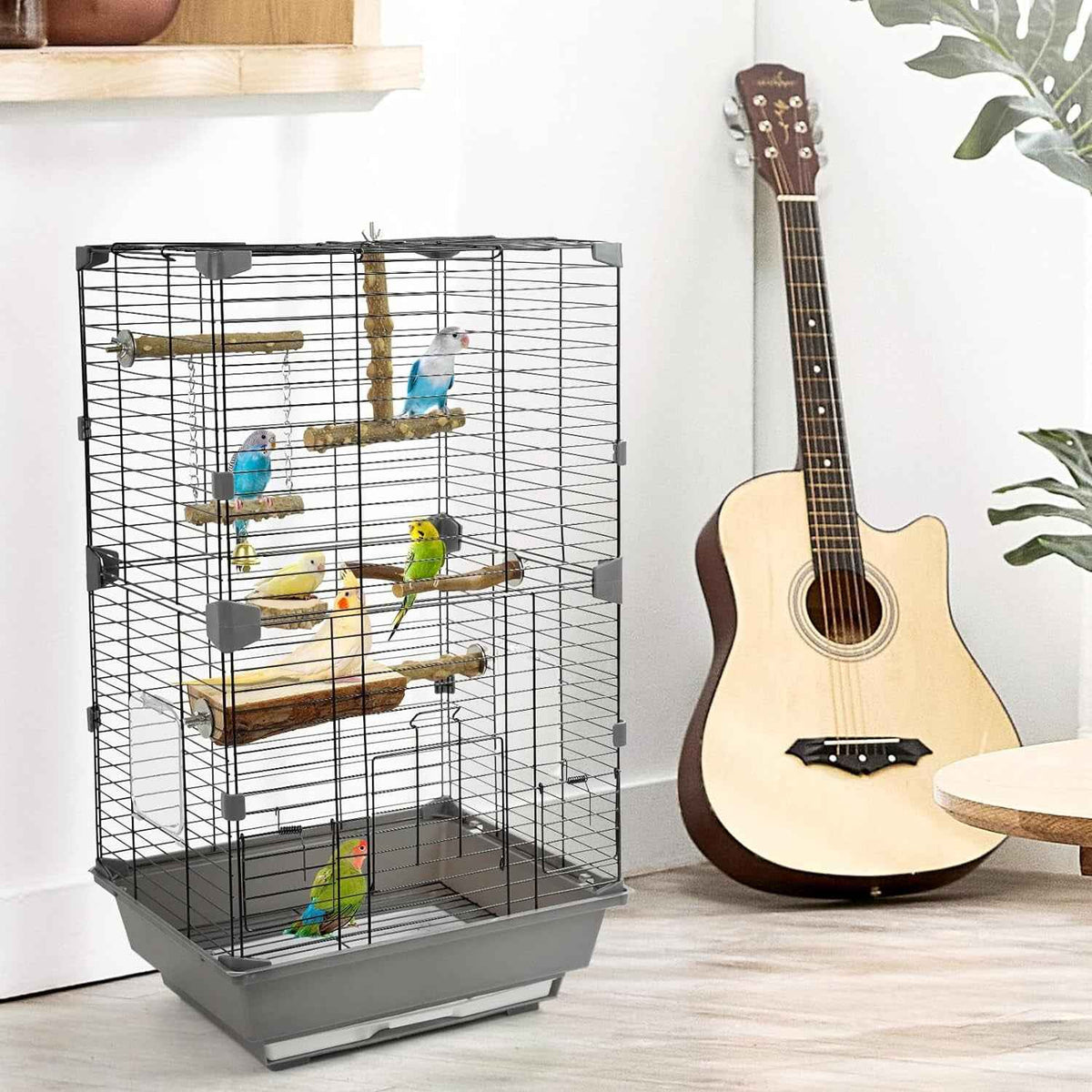 A large black wire bird cage with multiple wooden perches and a gray plastic tray, shown with several small colorful birds inside. The cage is placed in a room setting next to an acoustic guitar, with plants and a shelf visible in the background.
