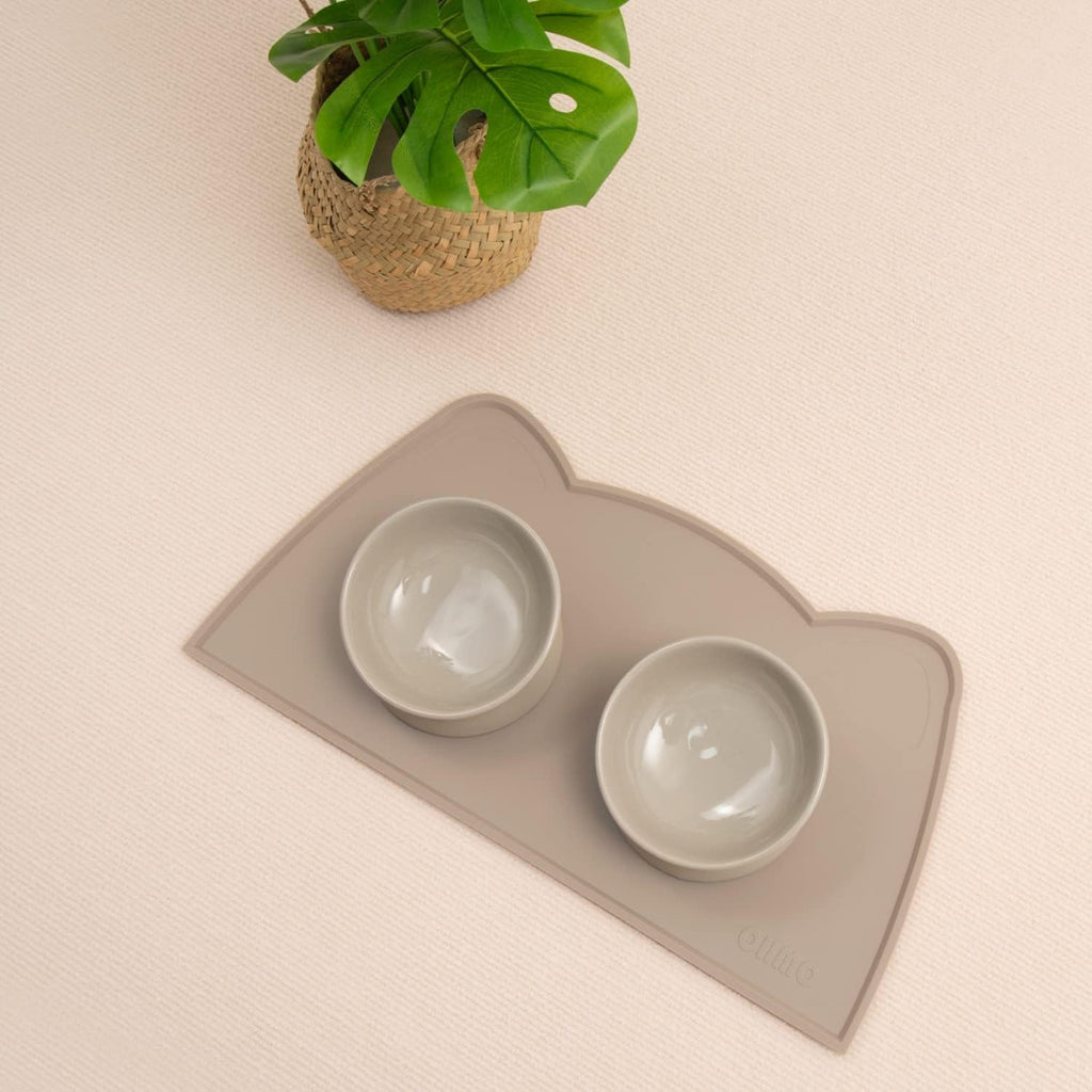 Cat-shaped mat with two bowls on a beige surface, accompanied by a plant.