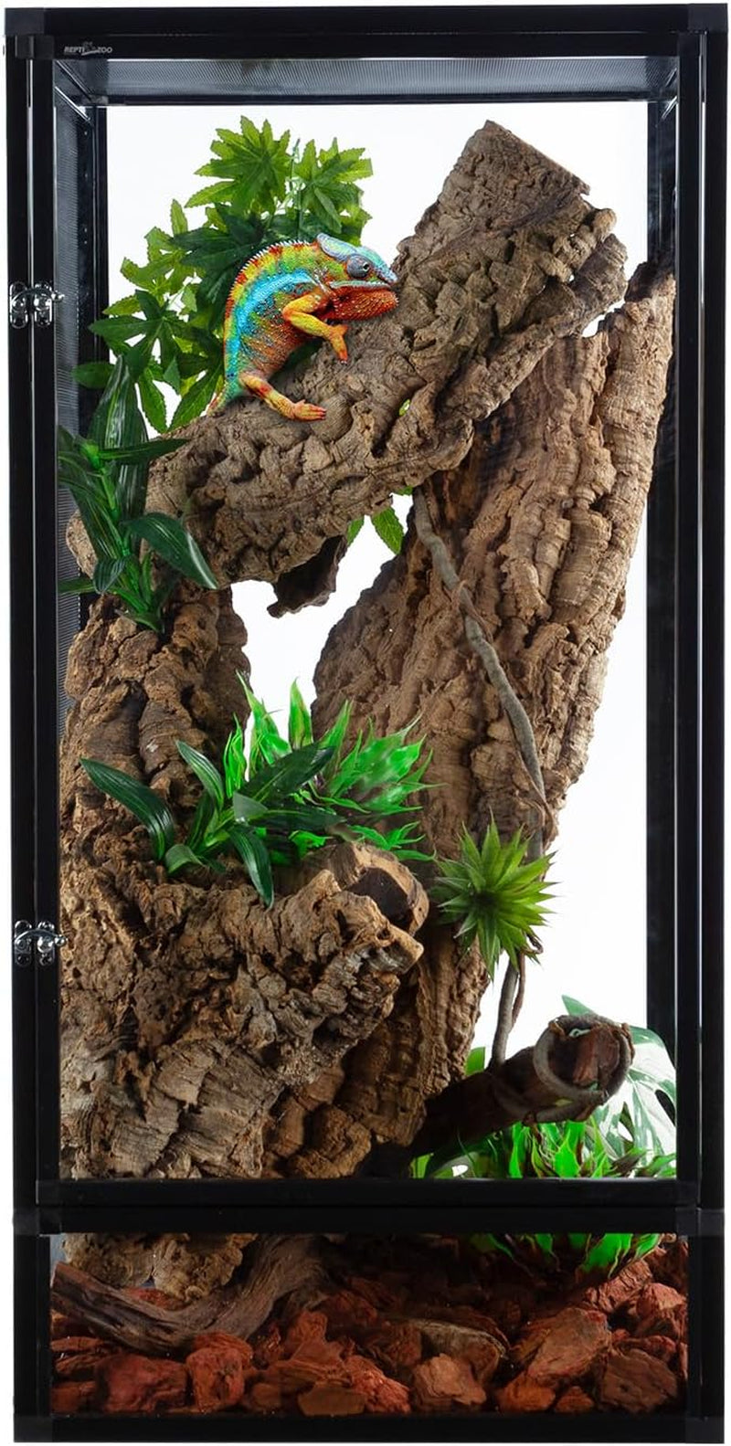 A live animal terrarium with a black frame, featuring a colorful chameleon perched on a large cork branch, artificial green plants, and reddish-brown wood chips at the bottom.
