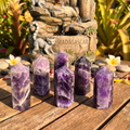 Six purple and white banded amethyst towers on a wooden surface with flowers and a decorative fountain behind them