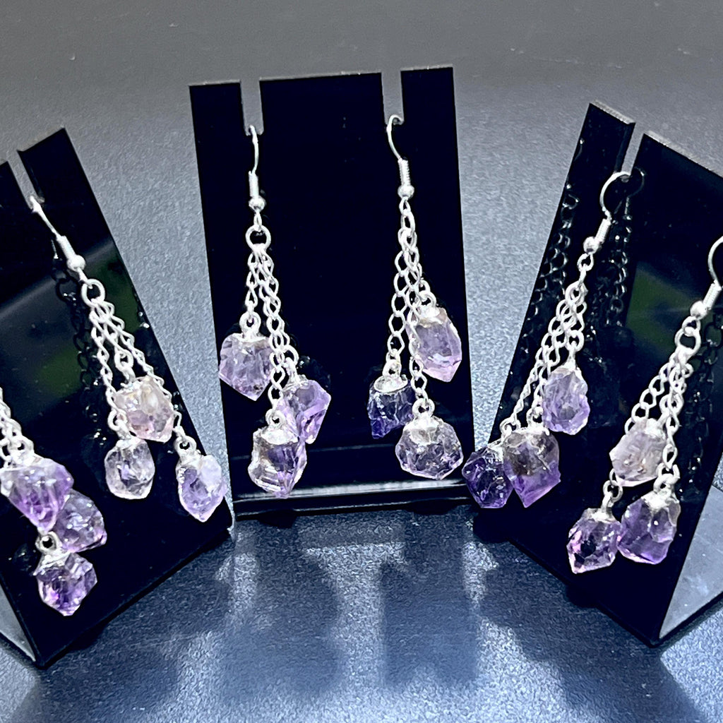  Amethyst triple crystal dangle earrings with silver-plated chains displayed on three black stands.