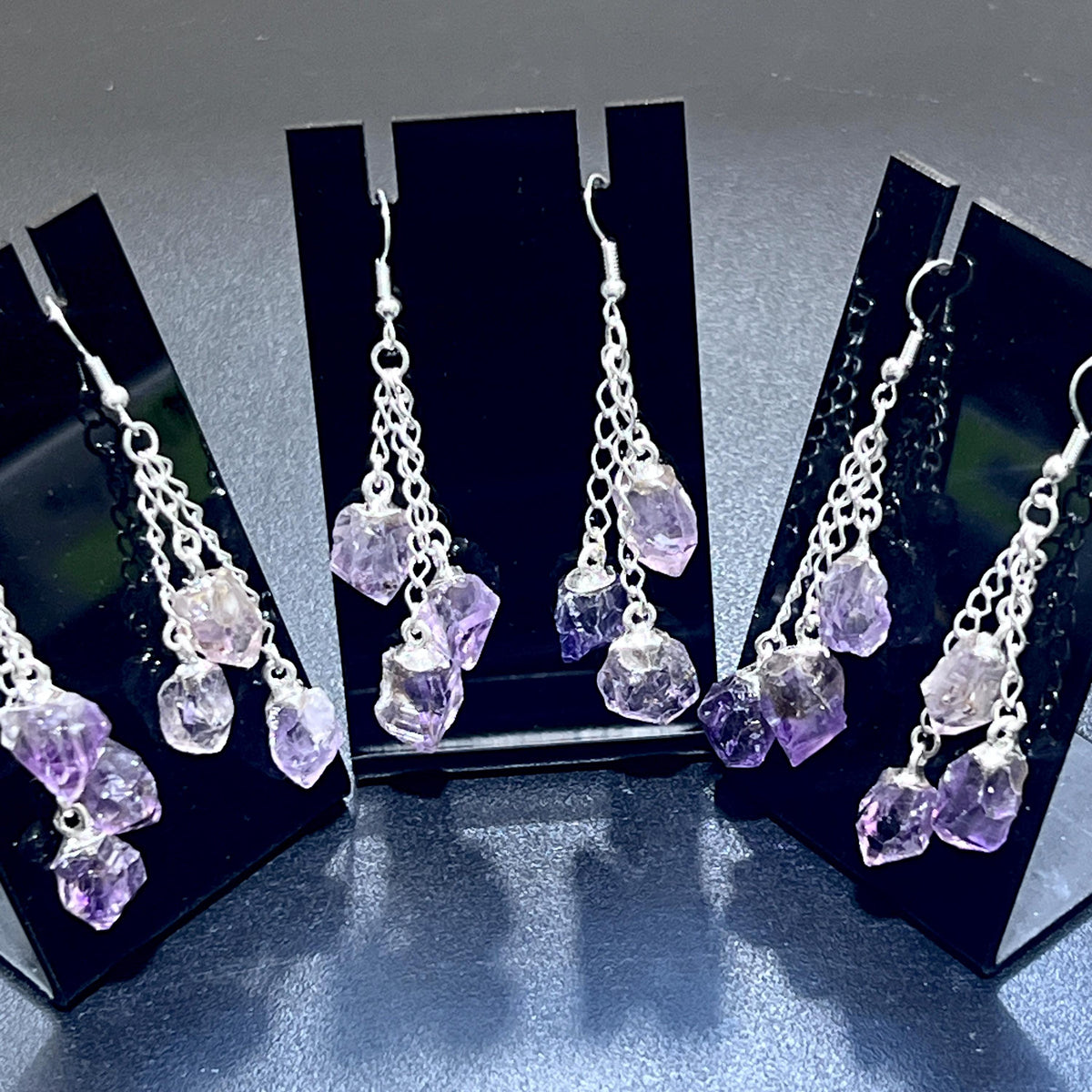  Amethyst triple crystal dangle earrings with silver-plated chains displayed on three black stands.