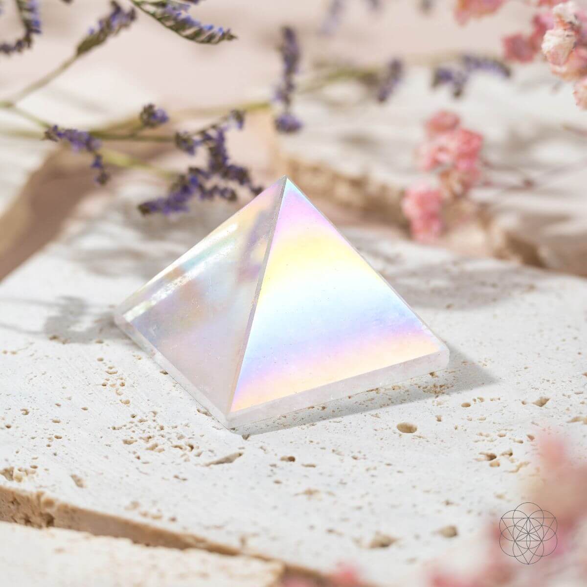 Pale Angel Aura quartz pyramid reflecting pastel rainbow colors on a cream stone slab beside flowers.

