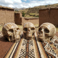 Three polished brown-banded aragonite skull carvings on a patterned textile with earthen buildings and hills behind them.