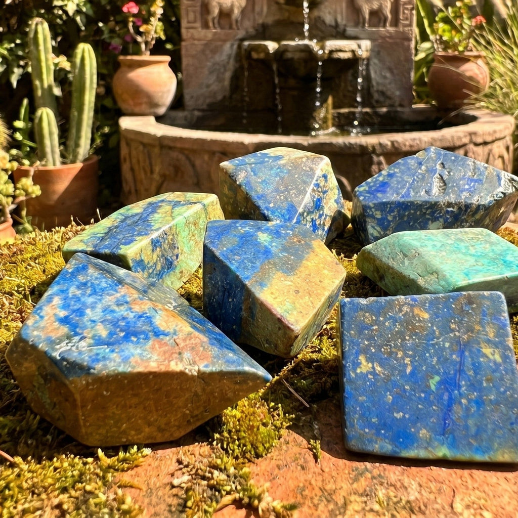 Blue and green polished azurite and chrysocolla freeform stones displayed on moss near a garden fountain.