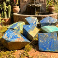 Blue and green polished azurite and chrysocolla freeform stones displayed on moss near a garden fountain.