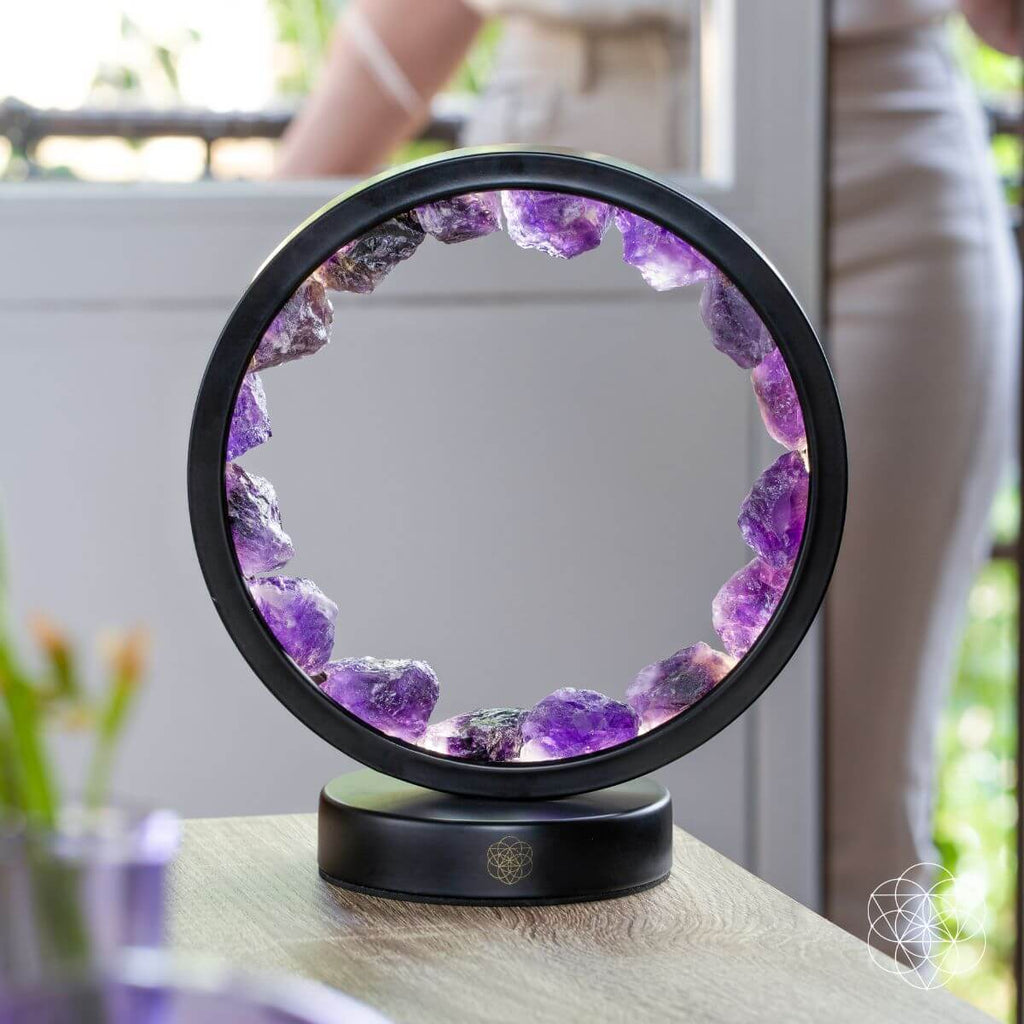 Black circular amethyst lamp showing rough purple stones in daylight on a wooden surface near a window.
