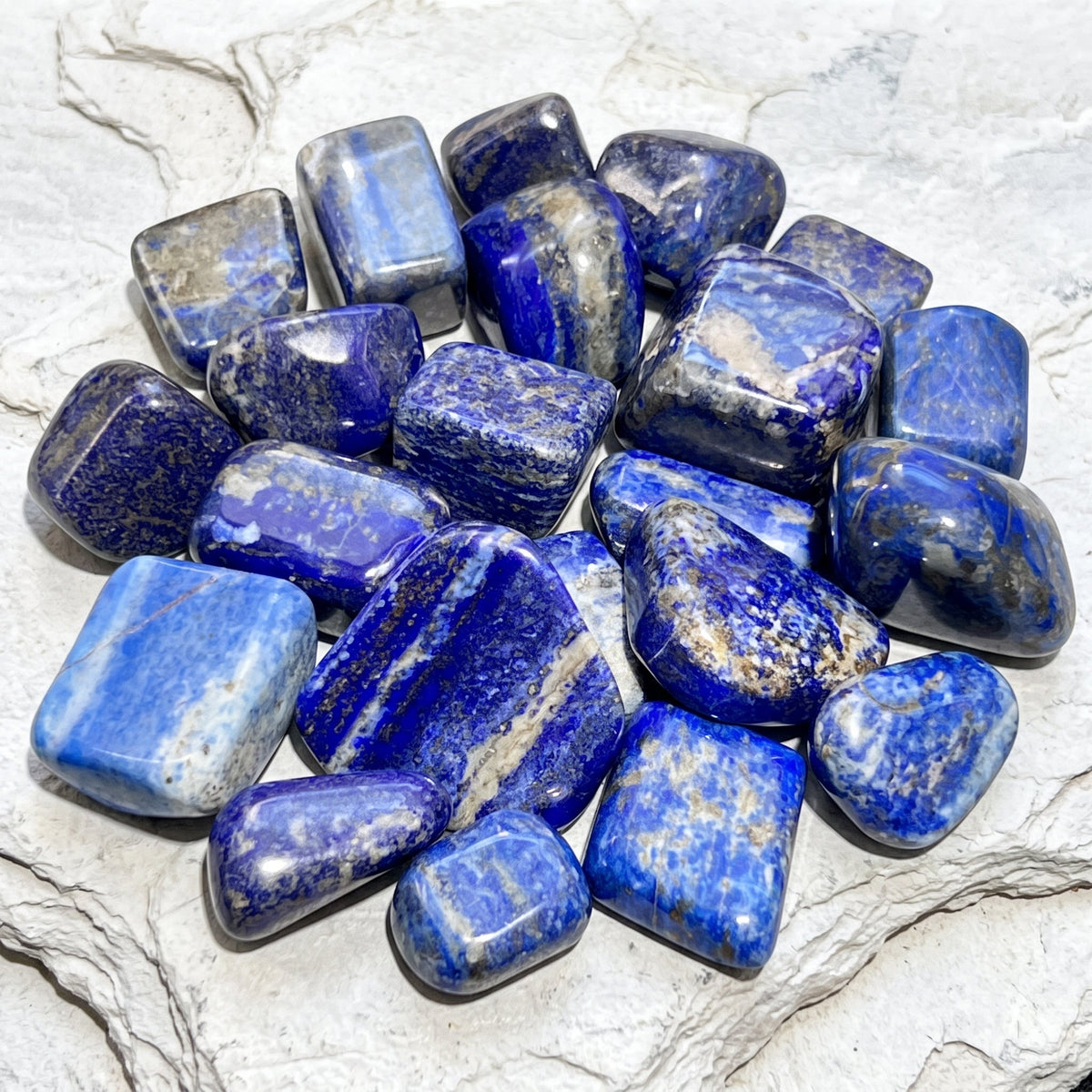 Blue lapis lazuli tumbled stones in varied rounded shapes displayed on a white veined stone surface.
