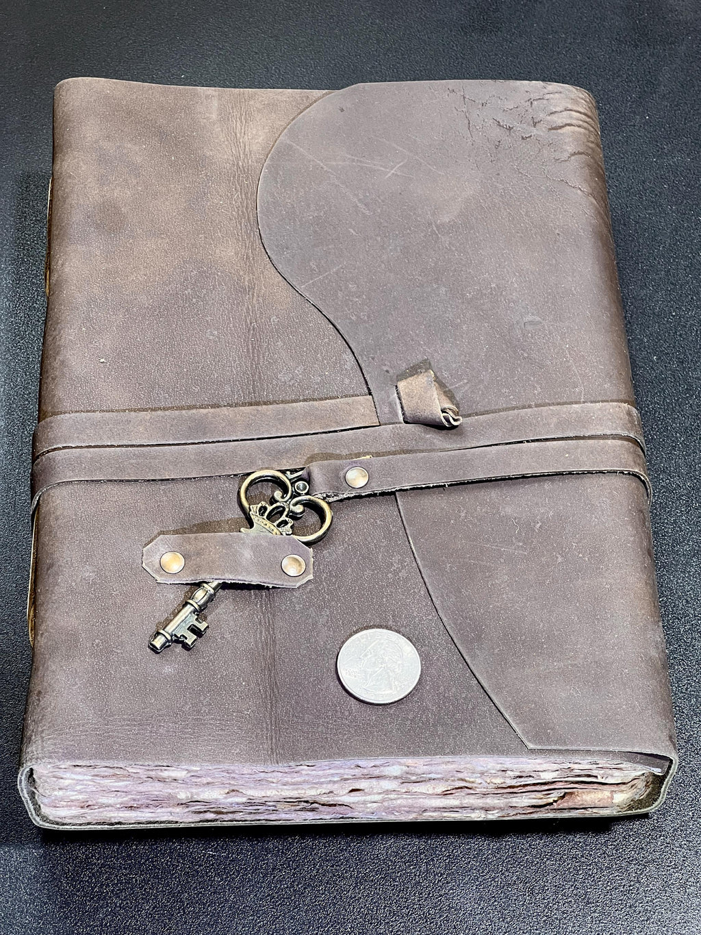 Brown leather sketchbook with curved cover flap, key detail and textured page edges beside a coin for scale