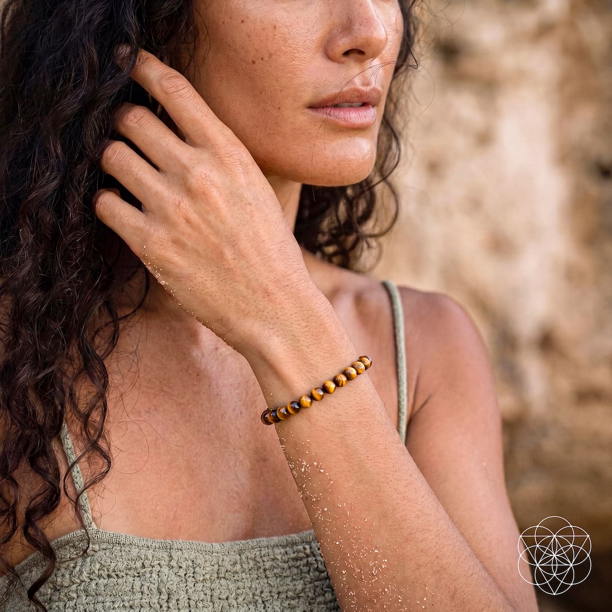 A woman outdoors wearing a beaded bracelet with dark brown and amber-orange beads. The photo is a close-up of her face, hand, and arm, with a blurred natural stone wall in the background.