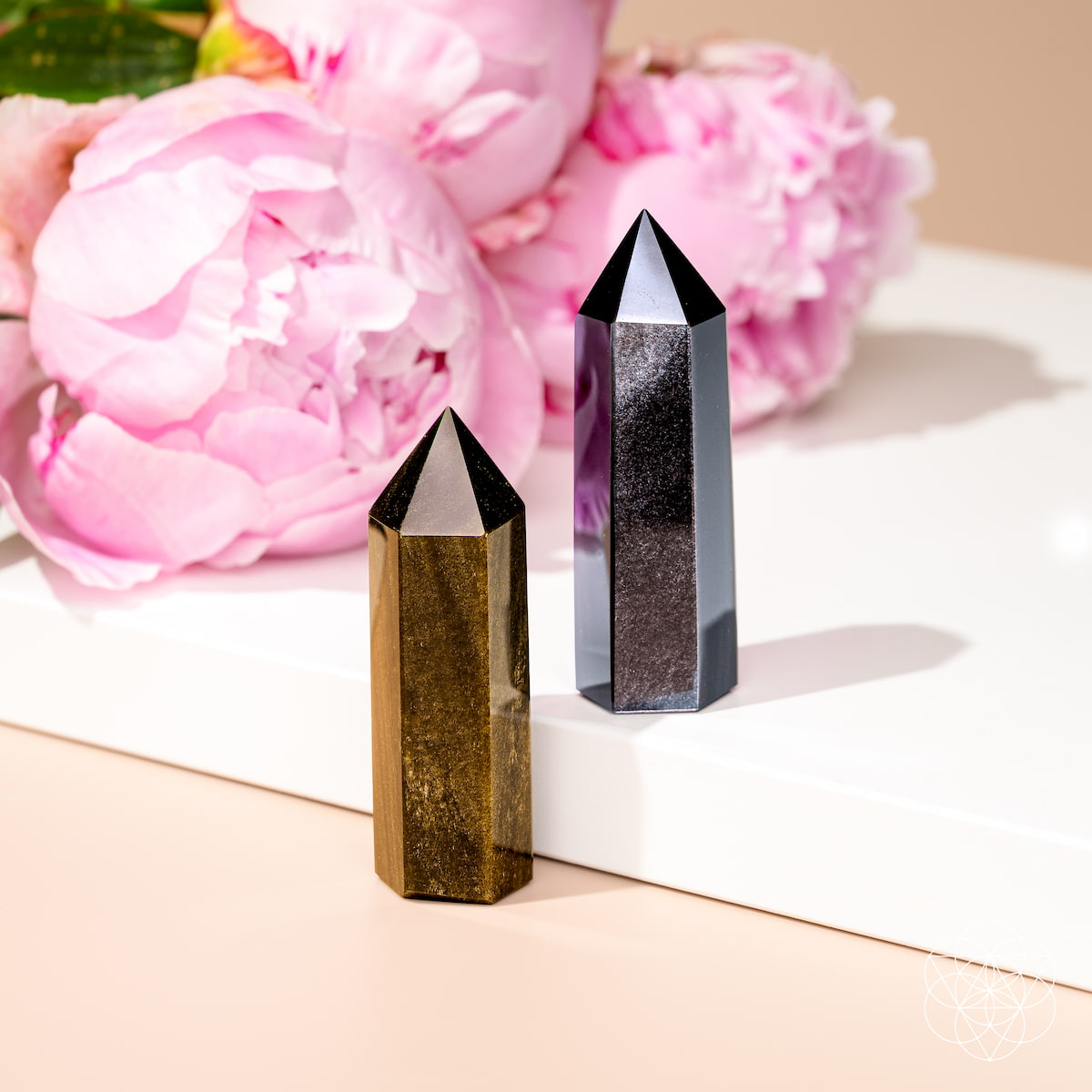 Gold sheen and silver sheen obsidian points standing beside pink flowers on a white display surface.
