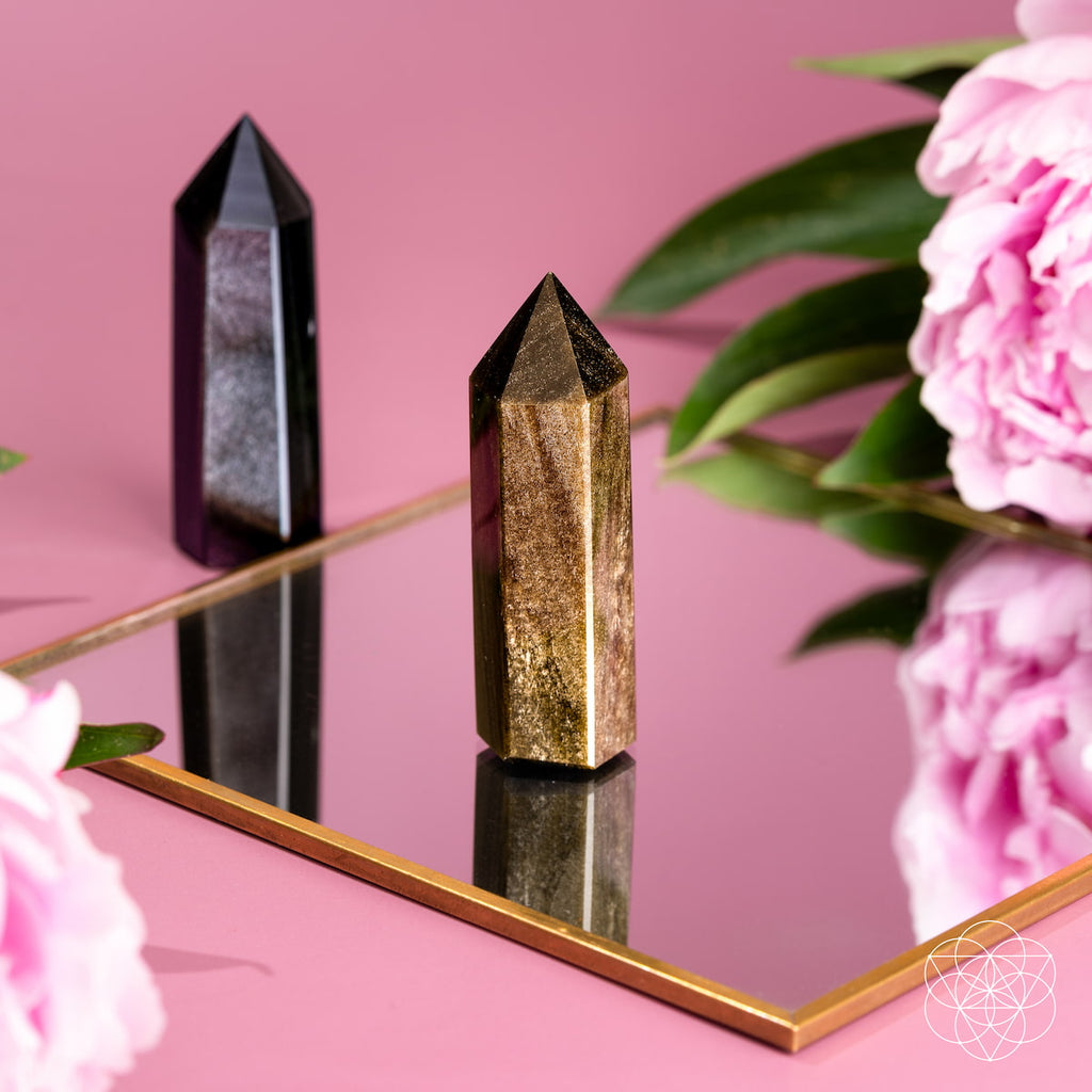 Gold sheen obsidian point standing on a gold-edged mirror, with a silver sheen point and pink flowers behind.
