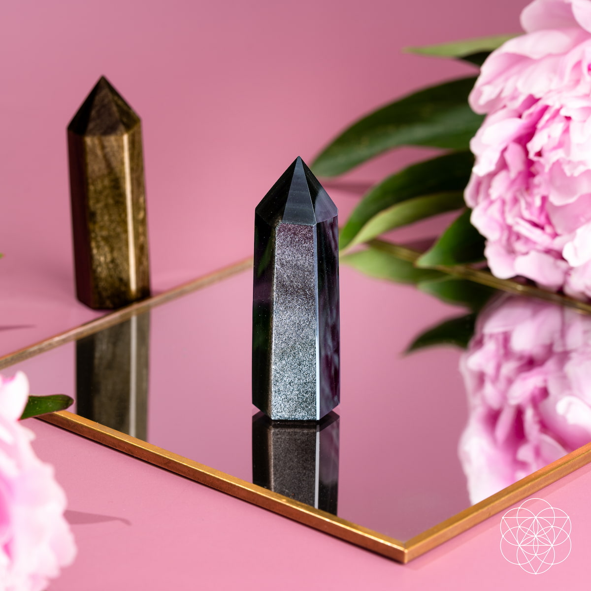 Silver sheen obsidian point on a gold-edged mirror with a gold sheen point and pink flowers in the background.
