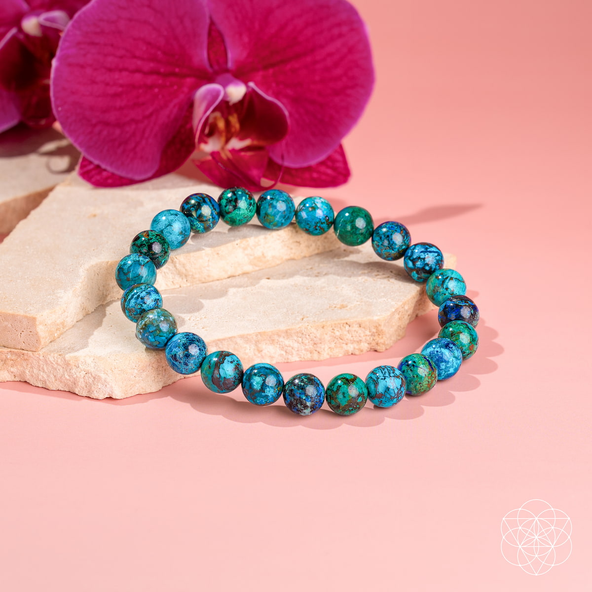 Blue-green chrysocolla bead bracelet displayed on cream stone slabs beside a magenta orchid.
