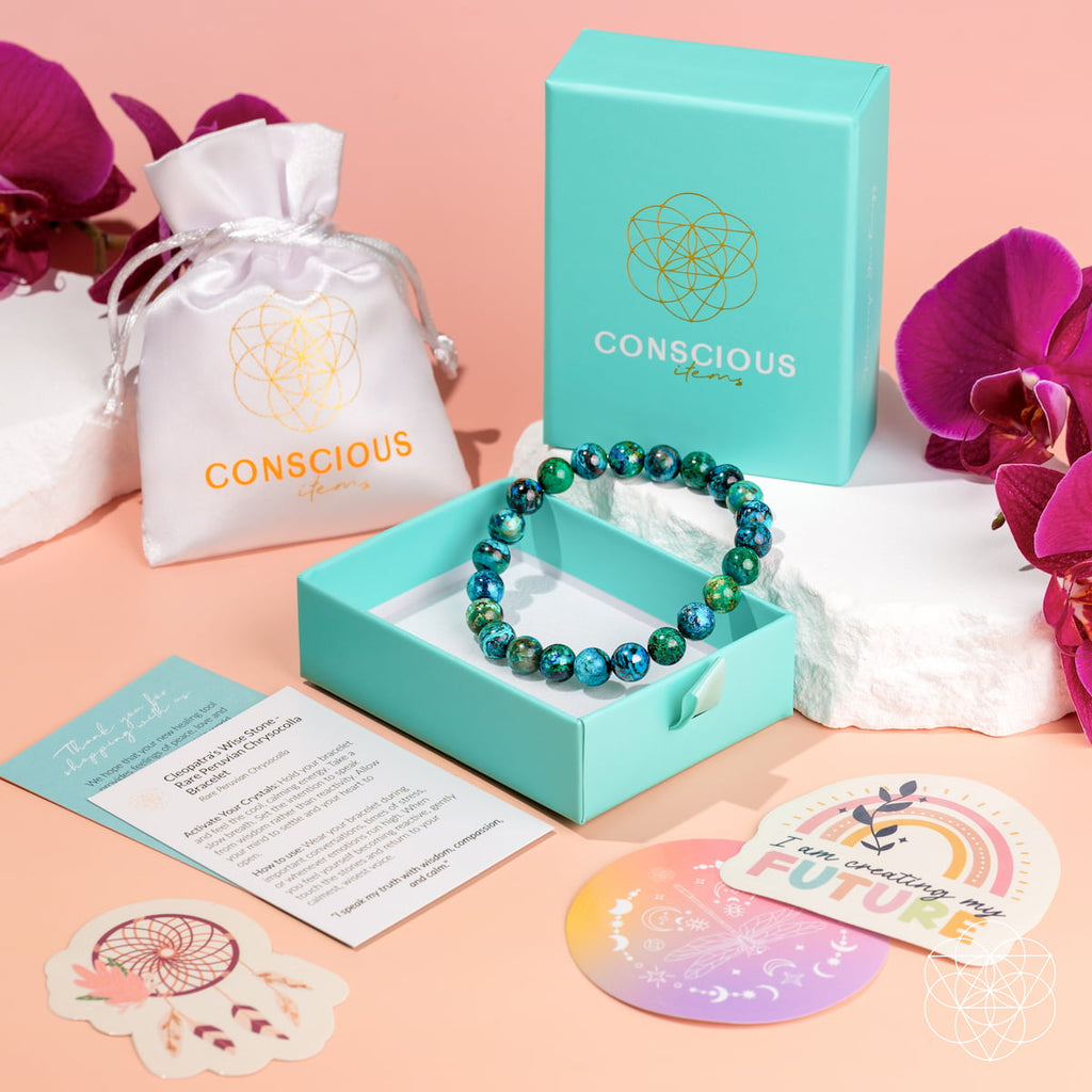 A beaded bracelet made of green and blue marbled beads displayed in an open turquoise box, with a matching turquoise branded box and white drawstring pouch behind it. Pink orchid flowers, white stones, and printed cards are arranged around it on a peach background.