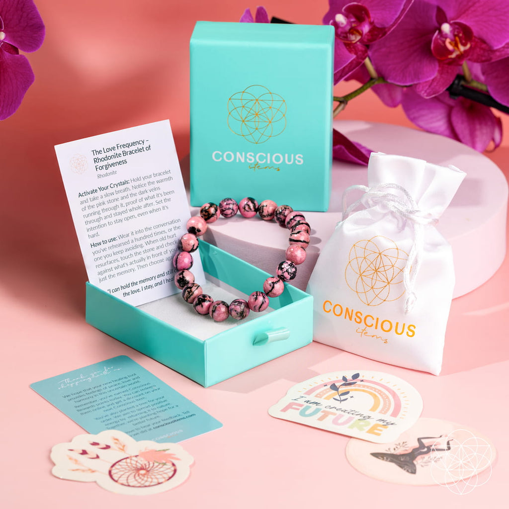 Rhodonite bracelet in a turquoise Conscious Items box beside a white pouch, cards, stickers, and orchids.
