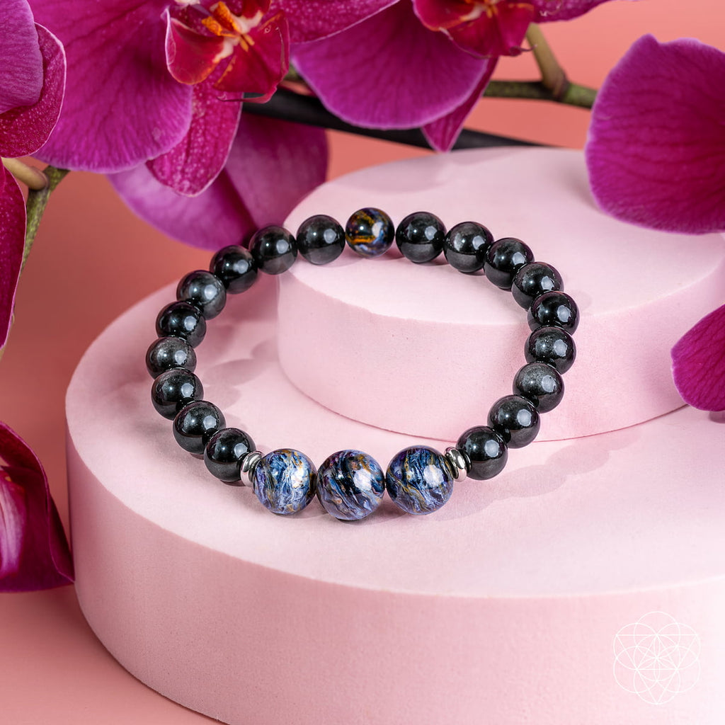 Pietersite and silver sheen obsidian bracelet on a pink display, with blue patterned focal beads and magenta orchids.
