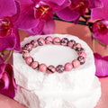 Pink rhodonite bead bracelet with dark veining on a white stone display surrounded by magenta orchids.
