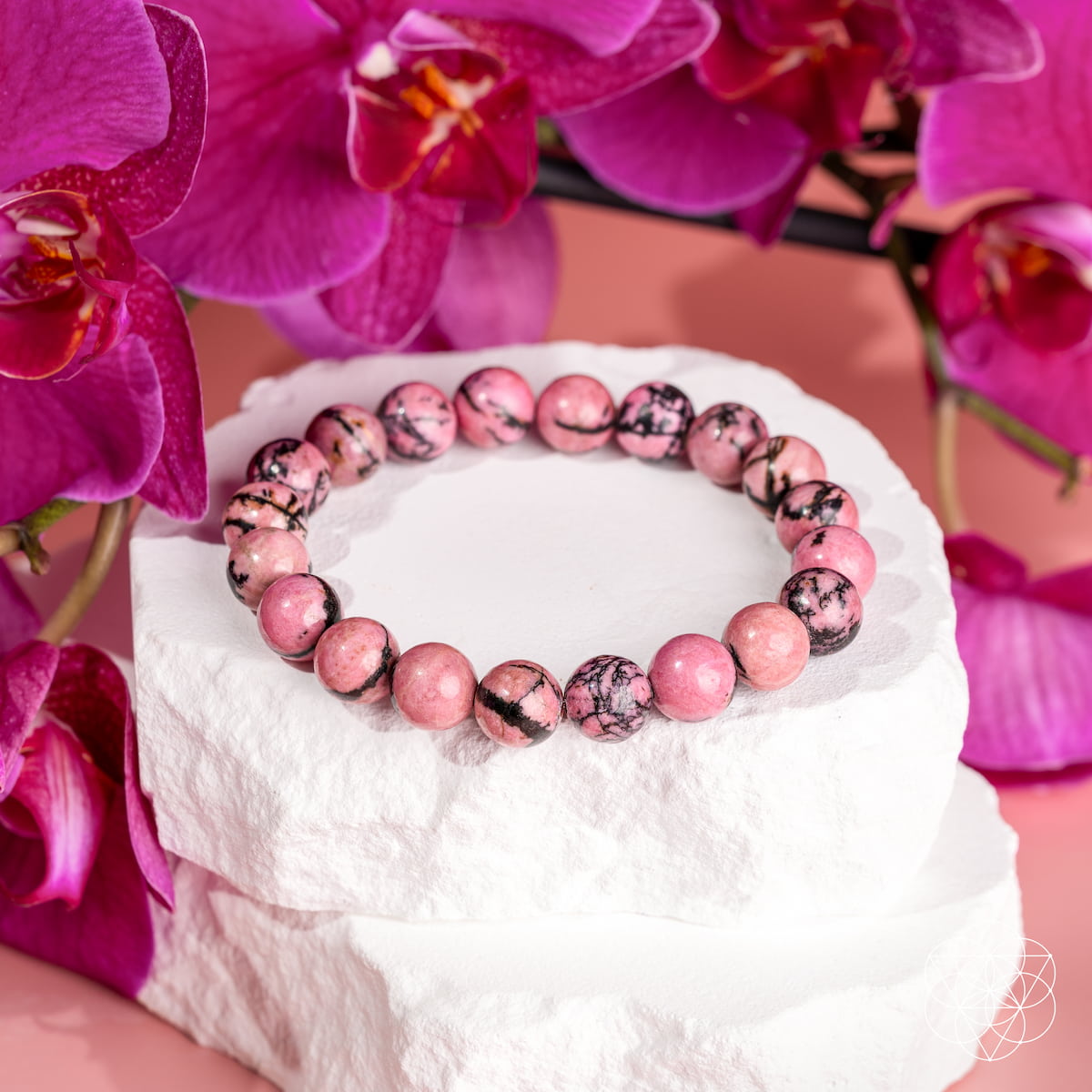 Pink rhodonite bead bracelet with dark veining on a white stone display surrounded by magenta orchids.
