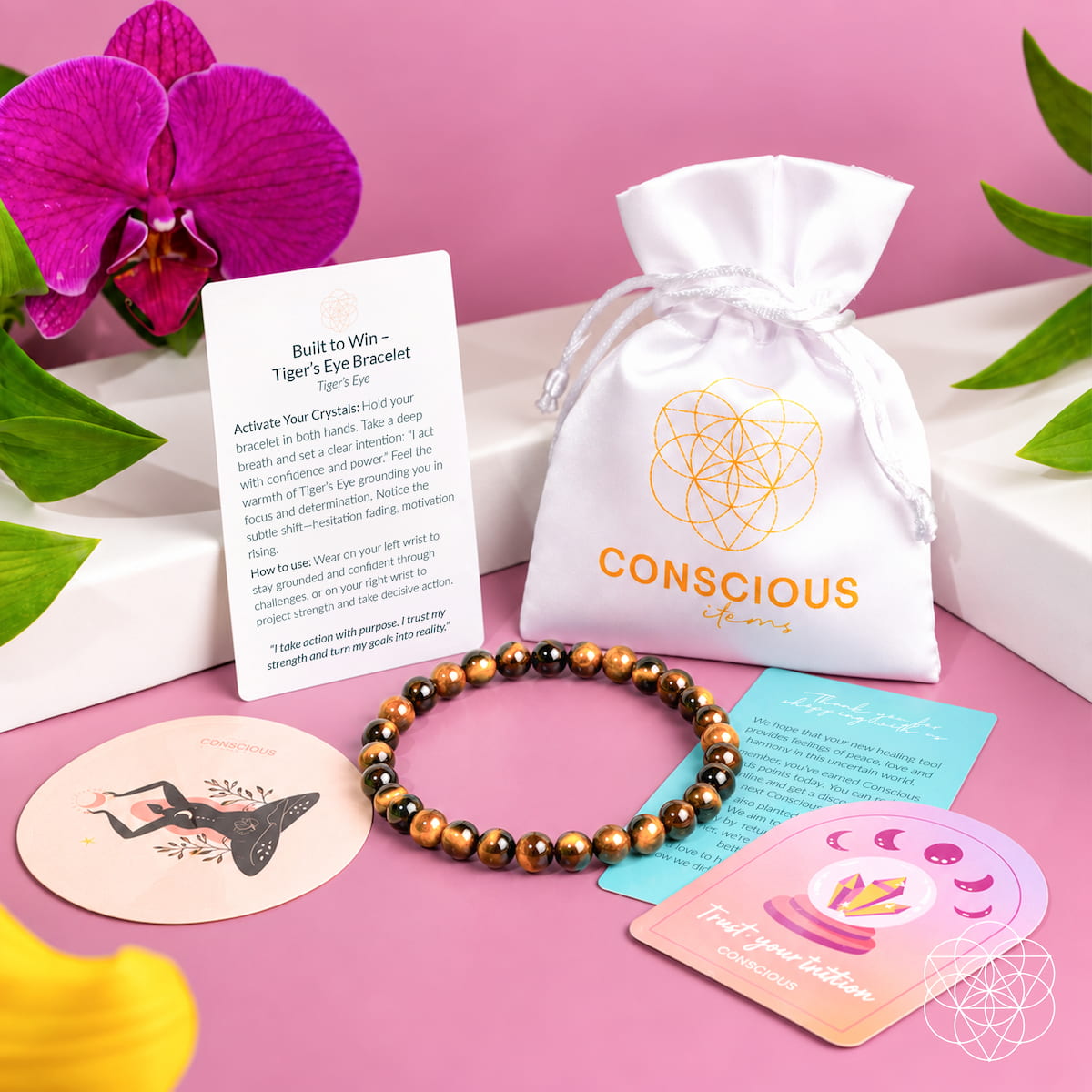 Tiger’s eye bracelet beside a white Conscious Items pouch, product card, and decorative cards on a pink surface.
