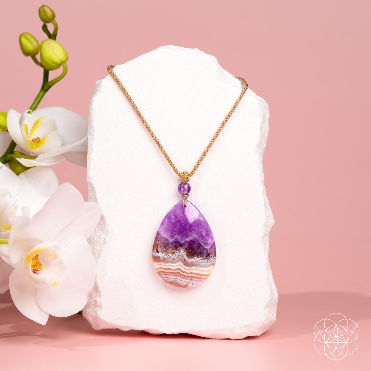 A teardrop-shaped amethyst geode pendant necklace with a rose gold chain, displayed on a white stone block with white orchids against a pink background.