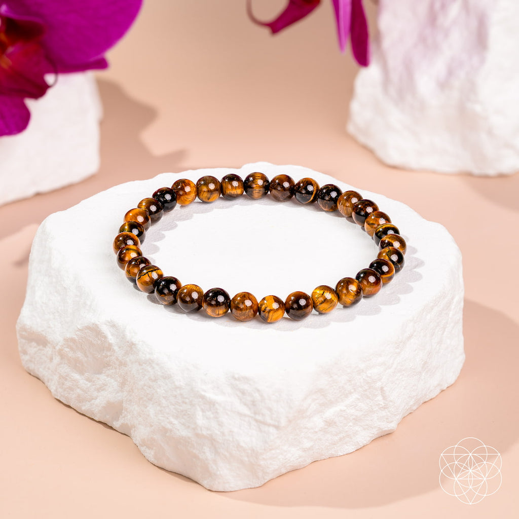 A beaded stretch bracelet made of polished tiger's eye beads in brown and black, displayed on a white stone pedestal against a soft peach background with blurred flowers.