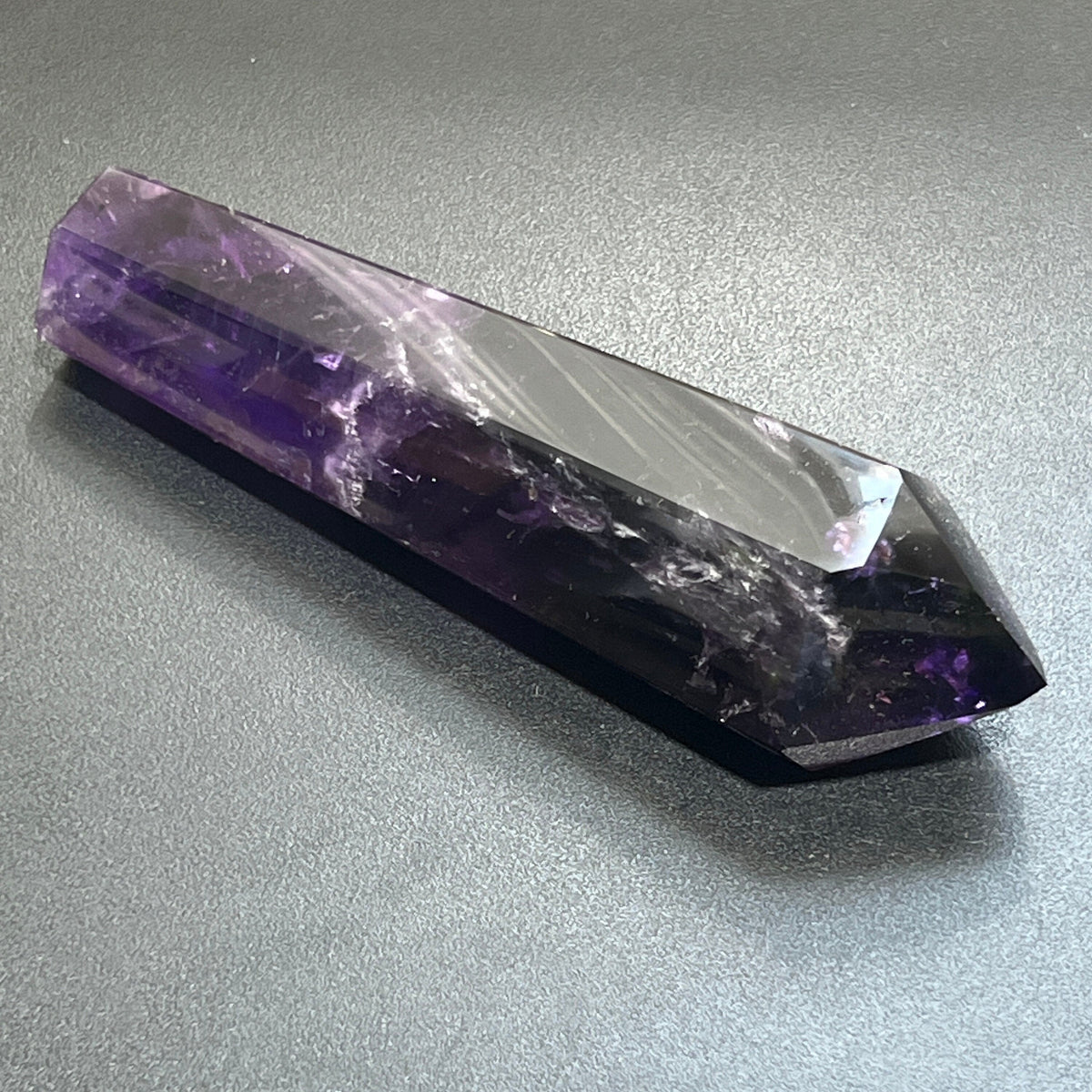 Dark amethyst tower lying diagonally on a gray surface, showing polished facets and pale internal markings