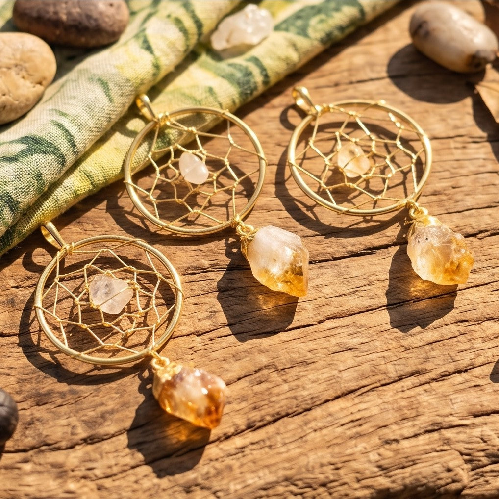 Three gold-plated dreamcatcher pendants with yellow crystal points displayed on sunlit wood beside patterned fabric.
