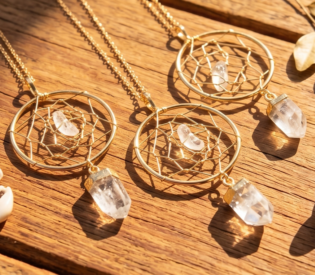 Three gold-plated dreamcatcher pendants with clear quartz points and chains arranged on sunlit wood.
