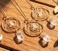 Three gold-plated dreamcatcher pendants with clear quartz points and chains arranged on sunlit wood.
