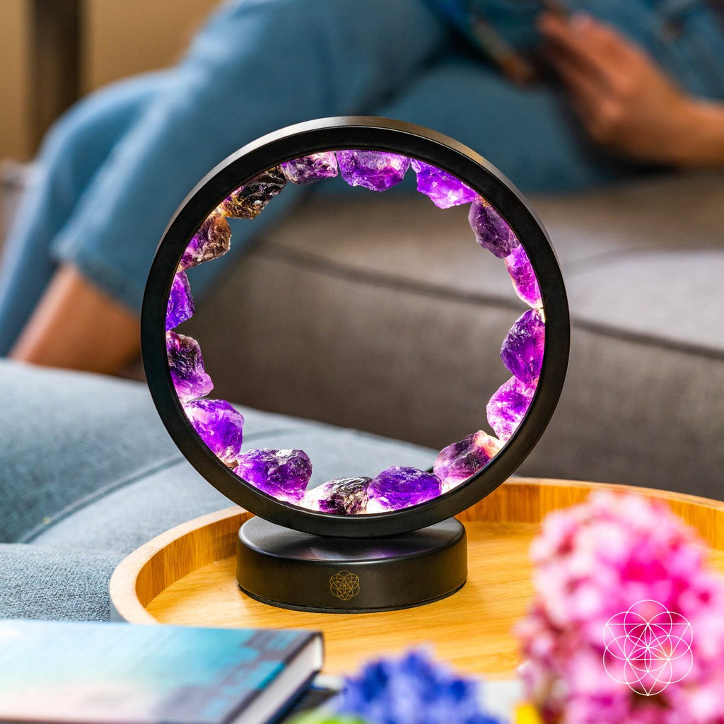 Glowing amethyst halo lamp on a round wooden tray beside a sofa, books, and pink flowers.

