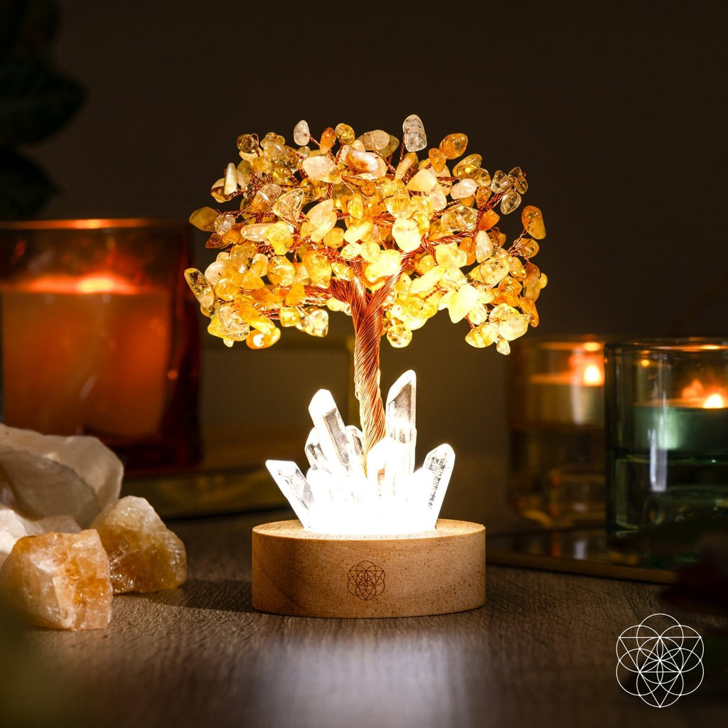 Glowing citrine crystal tree lamp with copper branches, clear quartz points, and a wooden base beside candles.
