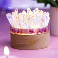 Glowing clear quartz point lamp with a pink tourmaline accent band and round wooden base on a pink surface.
