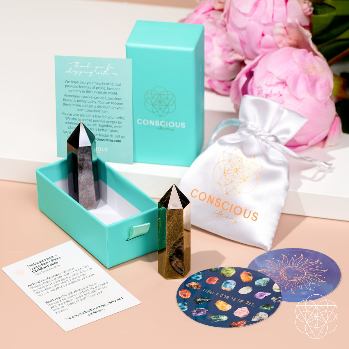 Gold and silver sheen obsidian points displayed with turquoise Conscious Items packaging, white pouch, and cards.

