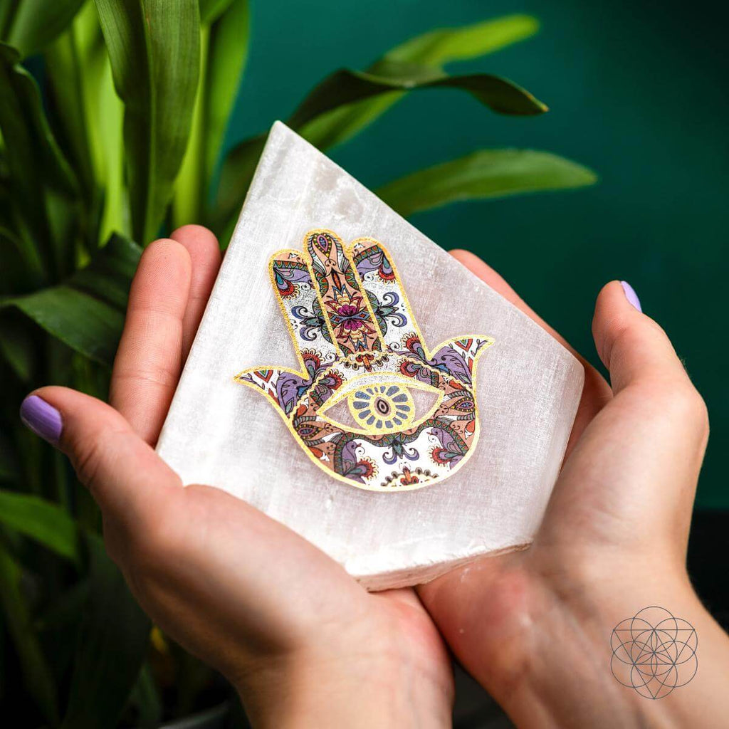 Person holding a pointed white selenite panel decorated with a multicolored Hamsa hand and central eye
