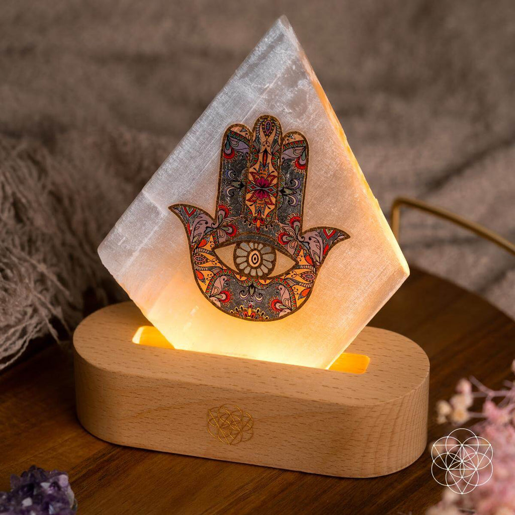 Angled close-up of a patterned Hamsa hand on a glowing selenite panel in a rounded wooden lamp base
