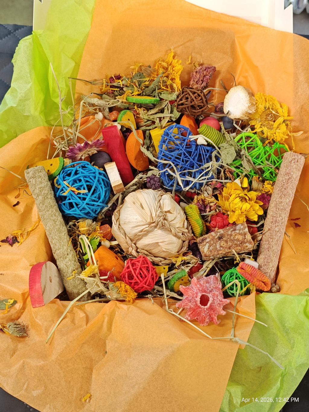 A colorful bird toy and treat box filled with various bird enrichment items including colorful woven balls, wooden blocks, dried herbs, and seeds, wrapped in orange paper and green tissue.