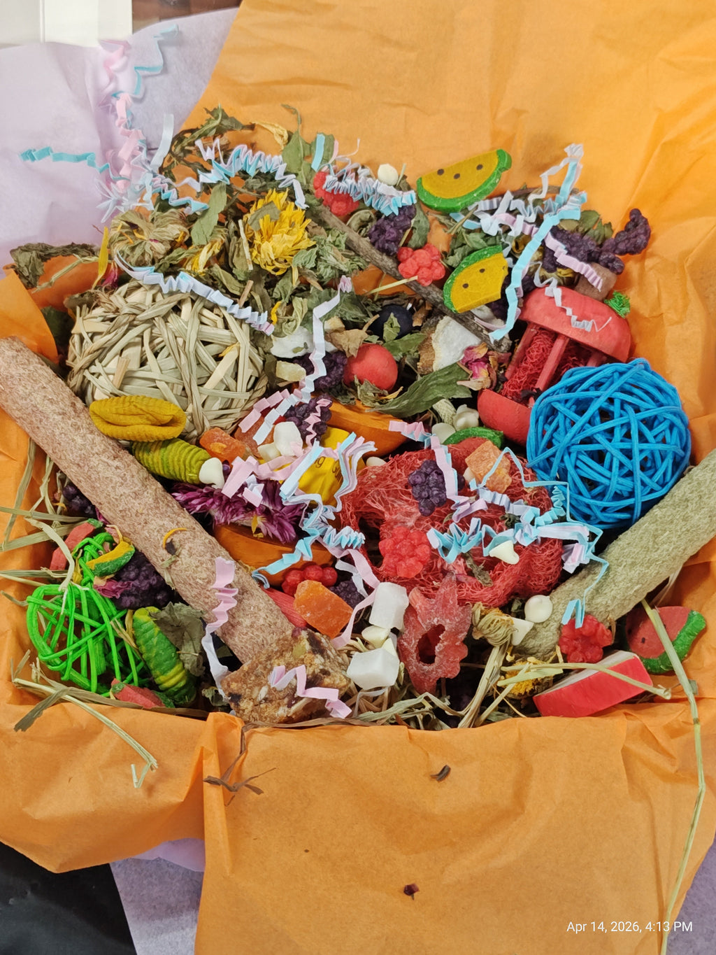 A colorful pet lick mat or chew toy bundle filled with dried herbs, treats, wooden sticks, and bright decorative pieces, wrapped in orange paper.
