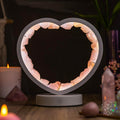 Illuminated rose quartz heart lamp with a pale frame and round base on a wooden table beside candles and crystals.
