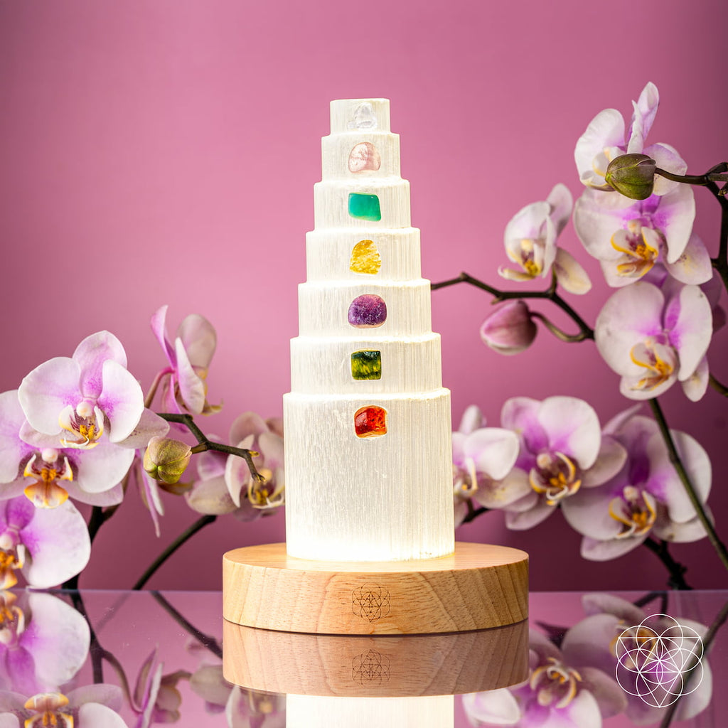  Illuminated selenite crystal tower on a wooden base surrounded by orchids against a pink background.
I