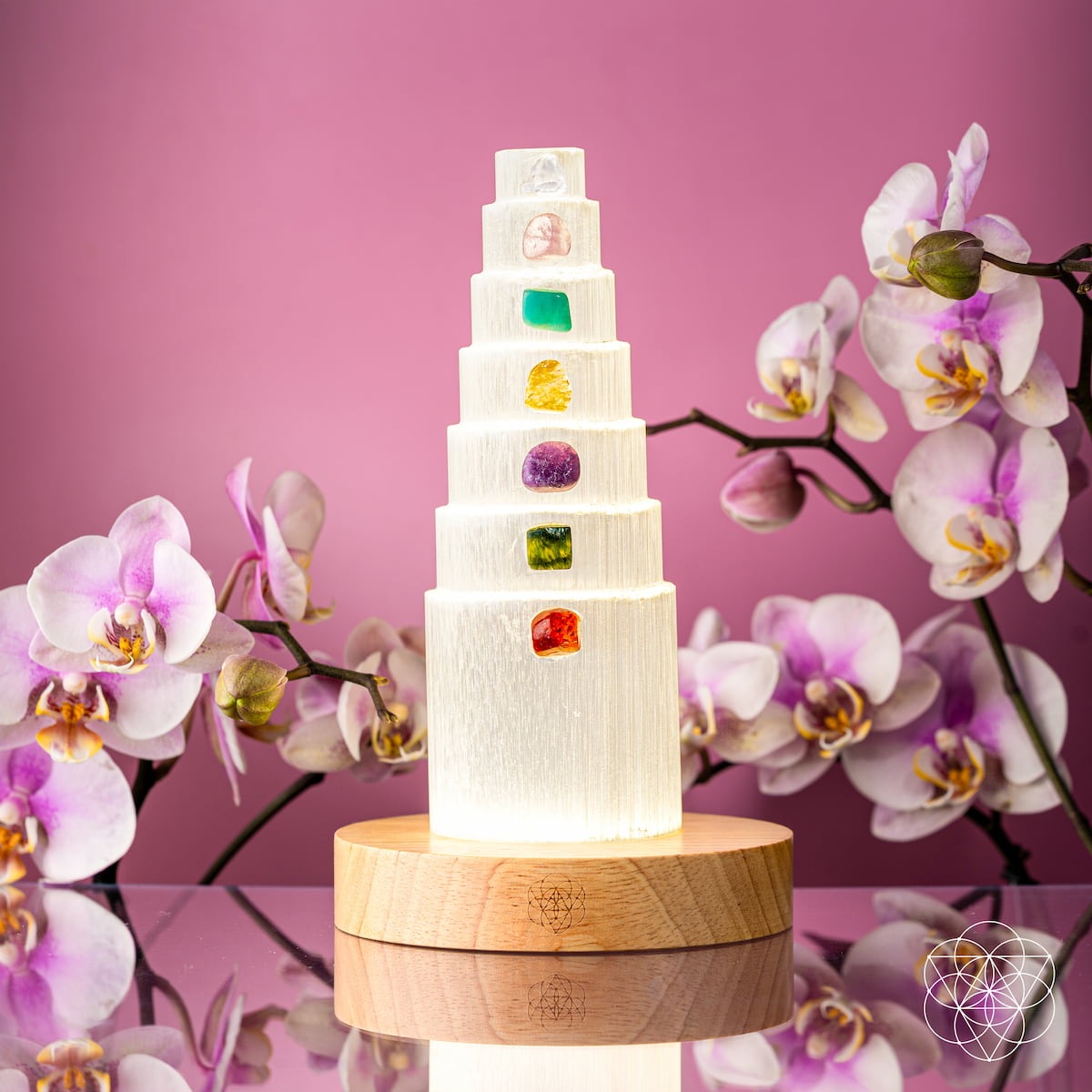  Illuminated selenite crystal tower on a wooden base surrounded by orchids against a pink background.
I