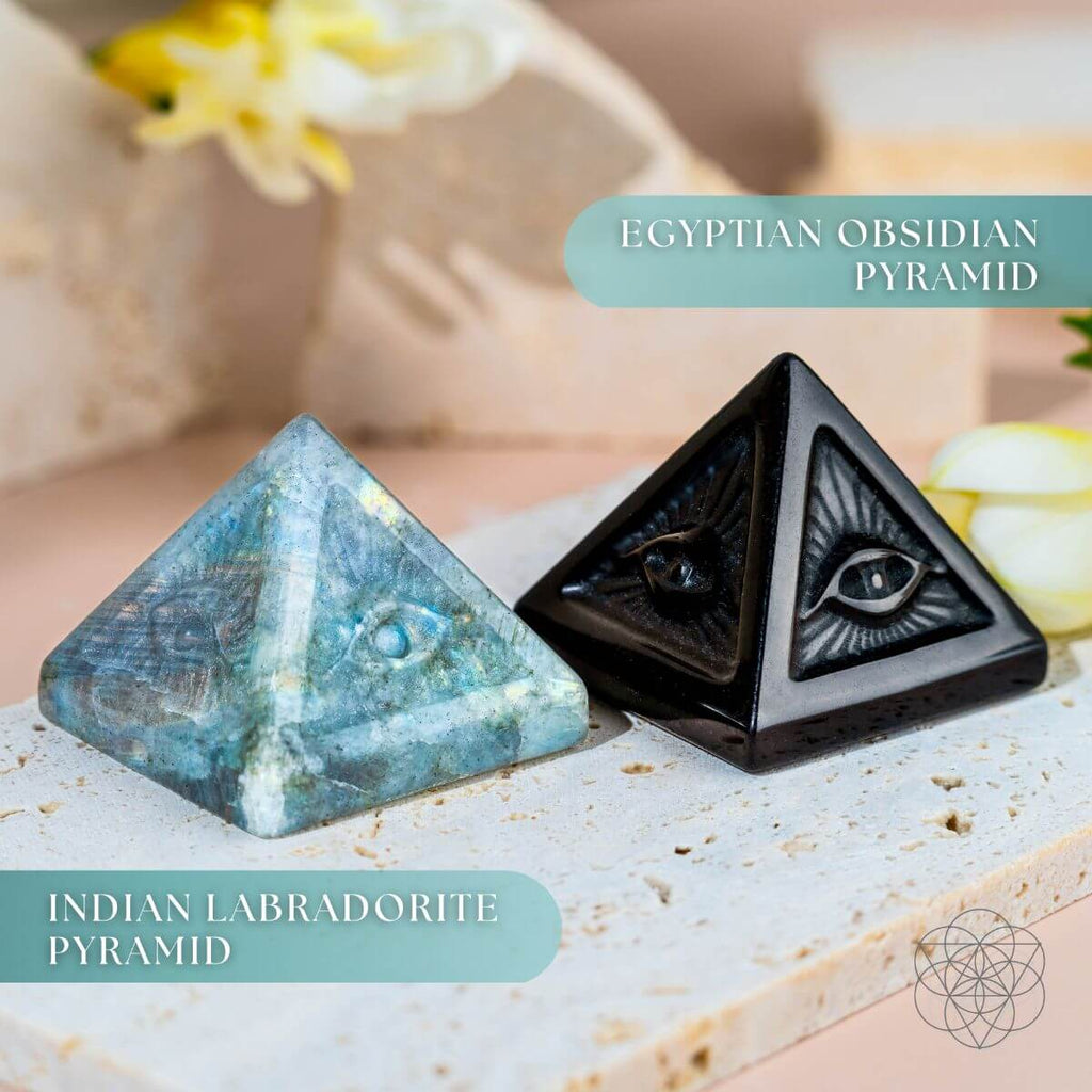 Labradorite and black obsidian pyramids with carved eye designs displayed together on pale stone slabs.
