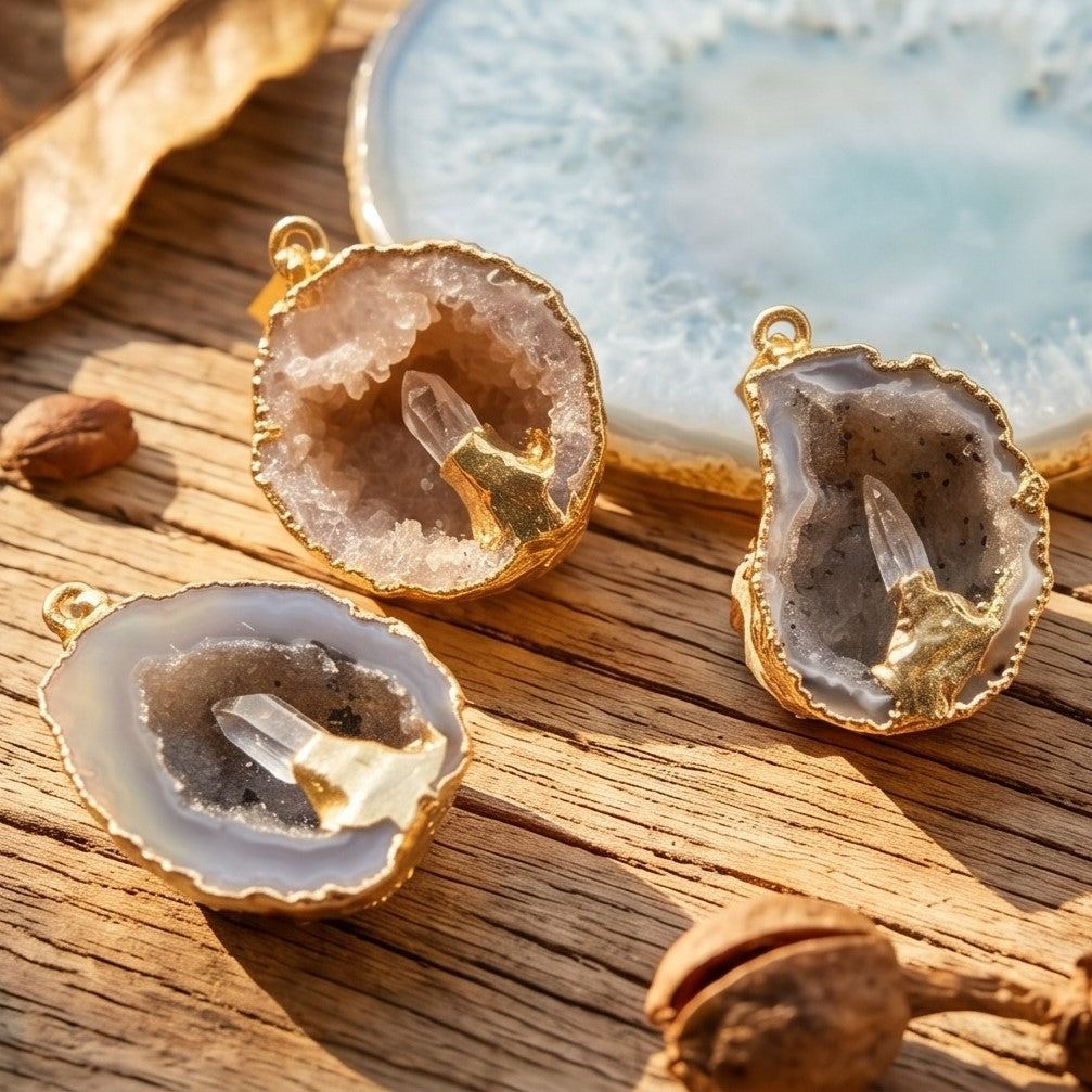 Three gold-edged Oco geode druzy pendants with clear quartz points displayed on sunlit wood.