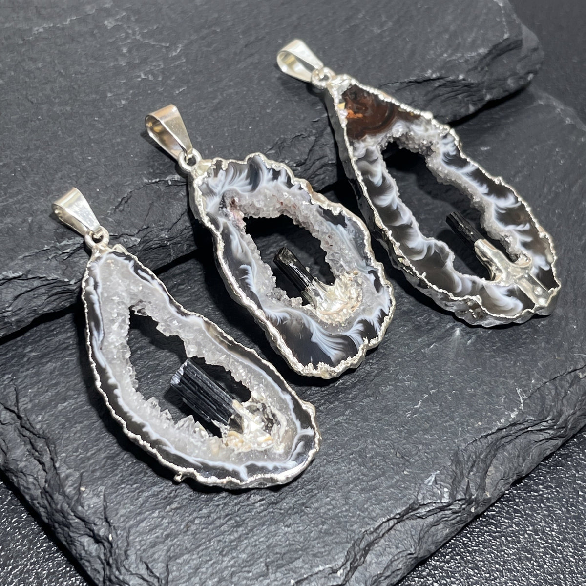 Three silver-plated Oco geode pendants with white druzy interiors, dark banding and black tourmaline accents on slate