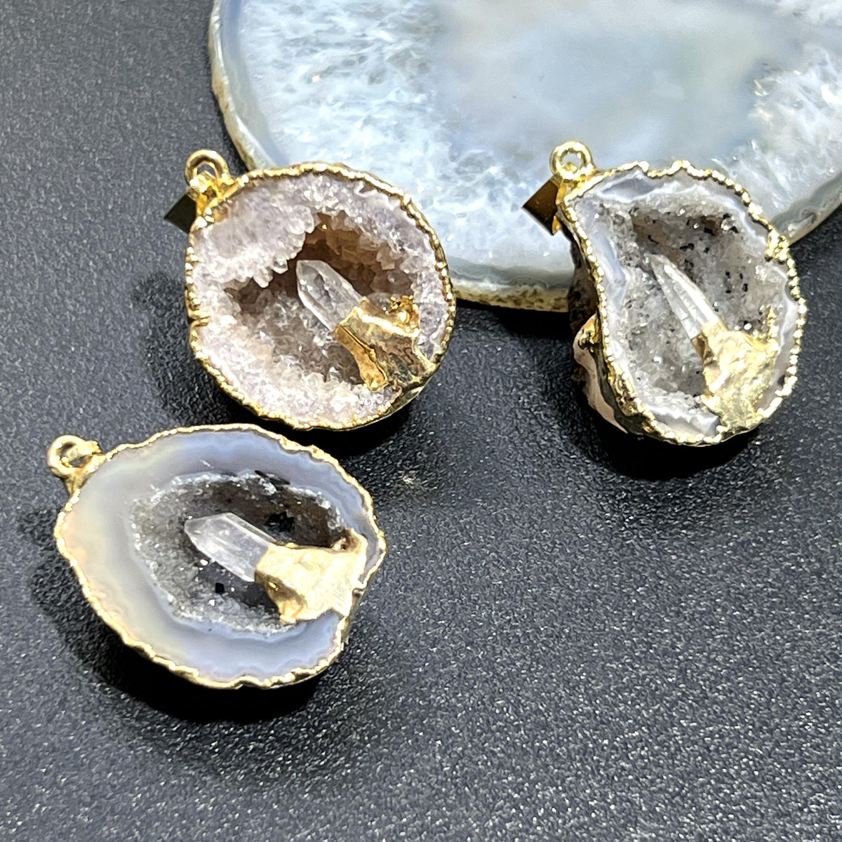 Oco geode pendants with crystal-lined openings, clear quartz points, and gold-plated edges on a dark surface.