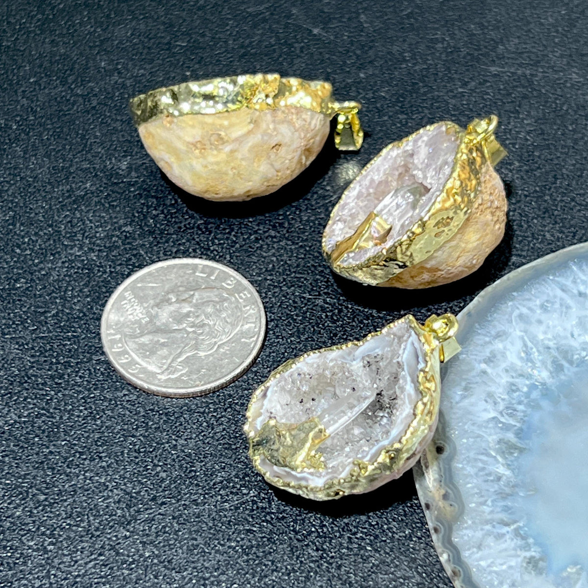 Side and front views of gold-plated Oco geode pendants showing rounded backs and quartz points beside a quarter.