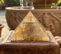 Golden yellow quartz resin pyramid with spiral details displayed on a carved wooden box near a fountain.