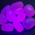 Pink mangano calcite tumbled stones fluorescing vivid pink and purple under UV light on a dark surface.